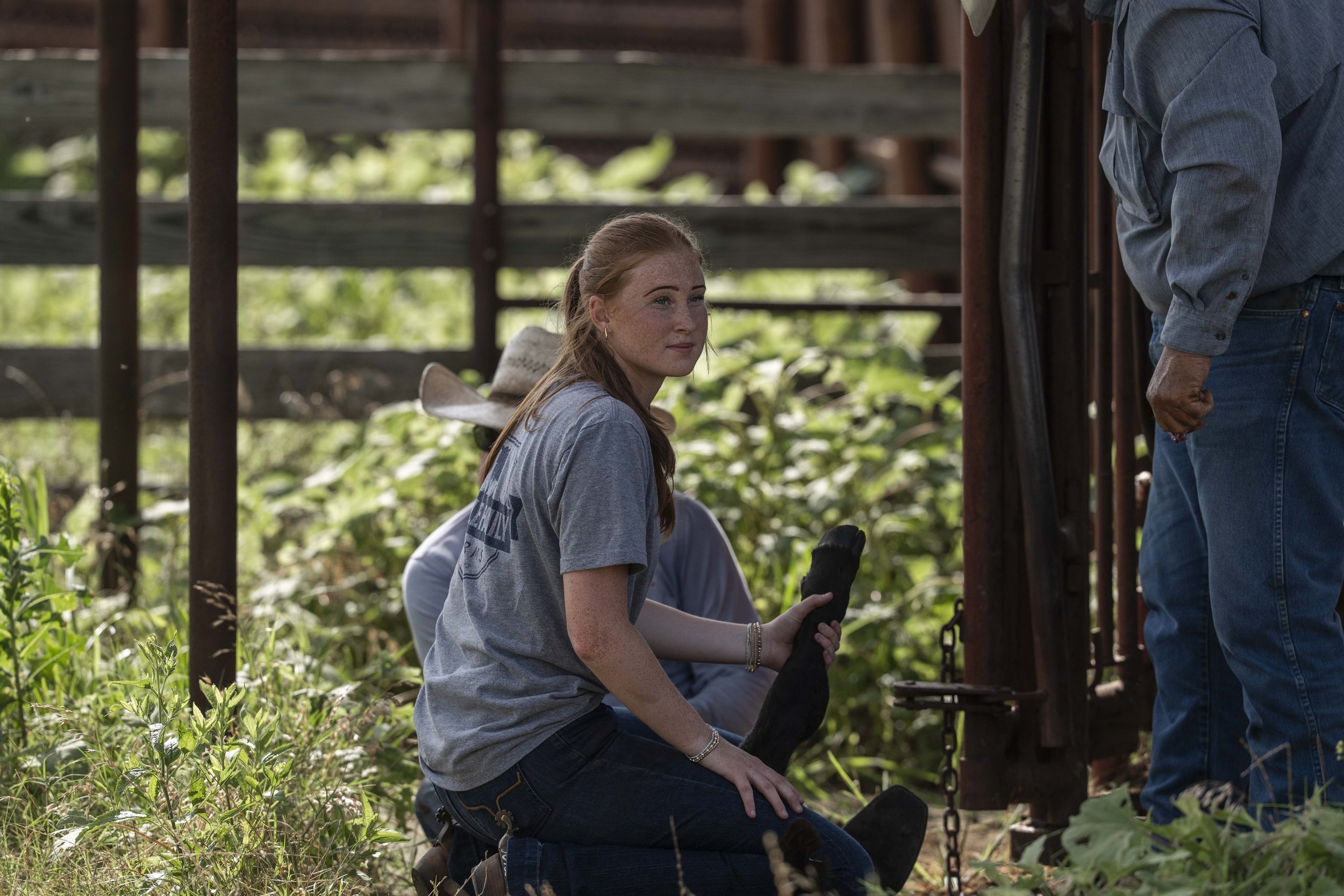 3322NOBLE Young woman in a gray t-shirt sits on the ground in a farm setting, assisting with livestock handling while two men are nearby. The background features greenery and a rustic fence, capturing a moment of agricultural teamwork.