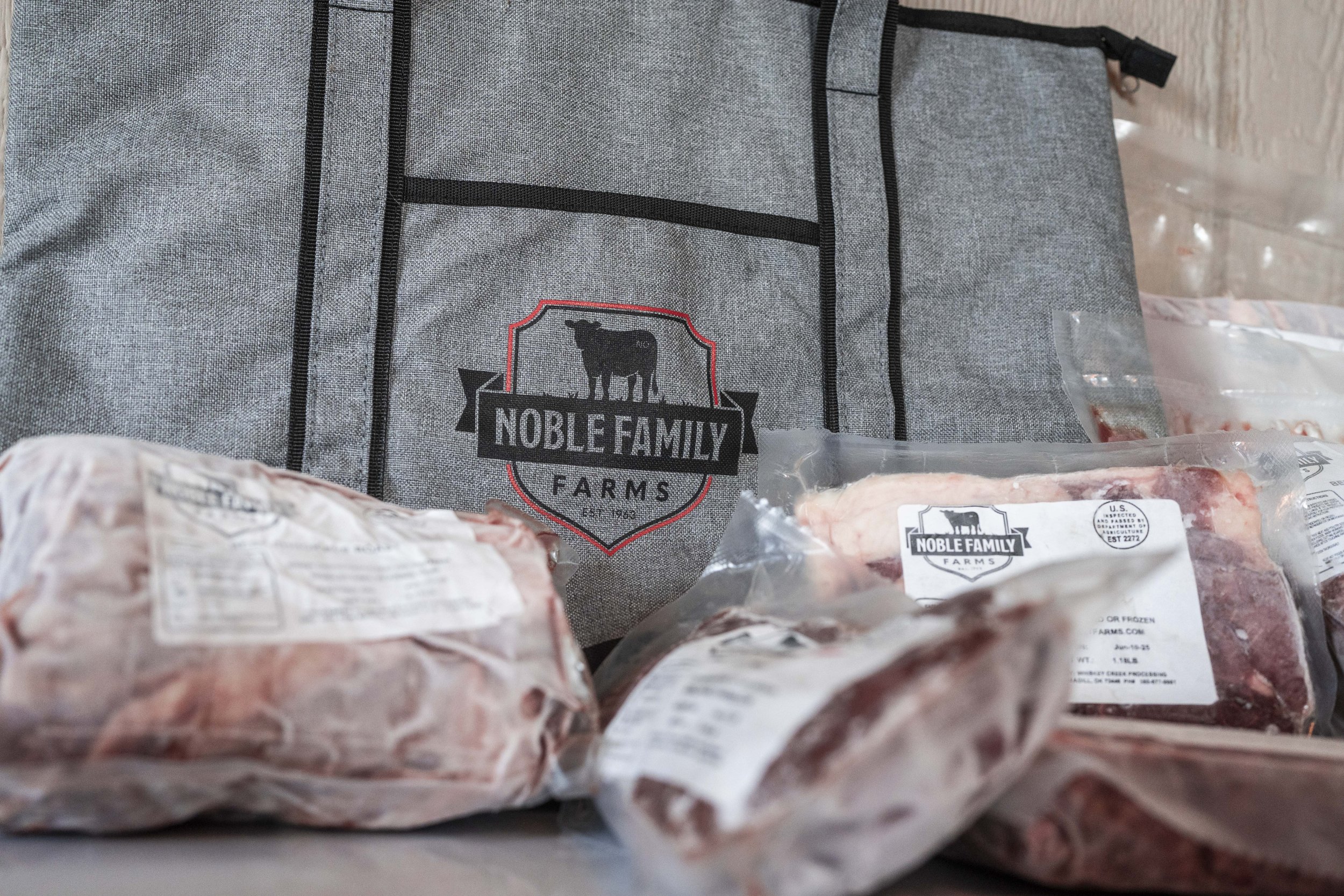 Noble Family Farms branded cooler bag with various packaged meats in the foreground, showcasing high-quality farm-fresh products.