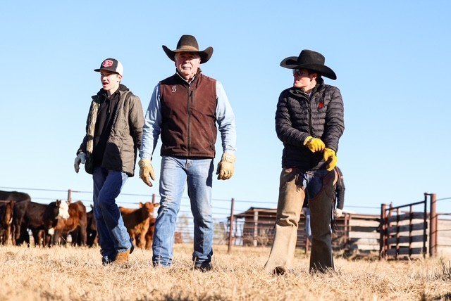 colt+denny+jace Three cowboys walking across a ranch field, dressed in winter attire and cowboy hats, with cattle visible in the background. They are engaged in ranch activities, showcasing a family-oriented farming lifestyle.